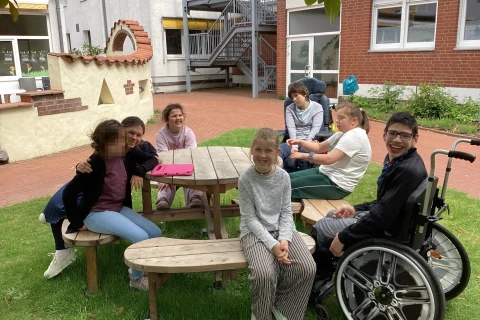 Kinder und Jugendliche, sechs sitzend und eines auf einem Holzpicknicktisch auf einem Rasenplatz, zwei in Rollst&uuml;hlen und eine mit verschwommenem Gesicht. Hintergrund mit Pflasterfl&auml;chen, modernen Geb&auml;udeteilen und einer Feuertreppe, flankiert von Pflanzen und B&auml;umen.
