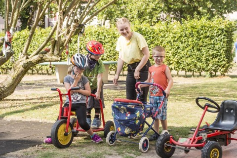 eine Betreuerin und 3 Kinder spielen draußen mit Spielfahrzeugen