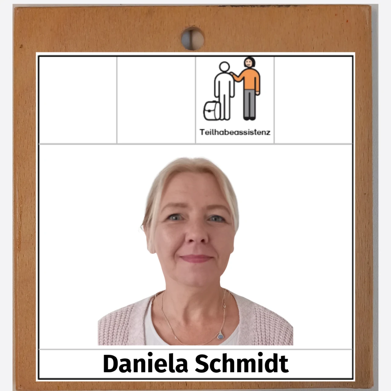 Portr&auml;tfoto einer blonden Frau mit neutralem Gesichtsausdruck vor einem wei&szlig;en Hintergrund. Oberhalb des Fotos ein Symbol mit zwei stilisierten Figuren, von denen eine einen Koffer tr&auml;gt. Daneben der Text: &bdquo;Teilhabeassistenz&ldquo;. Unterhalb des Fotos der Text: &bdquo;Daniela Schmidt&ldquo;. Das Bild ist in einem schlichten Holzrahmen eingefasst.