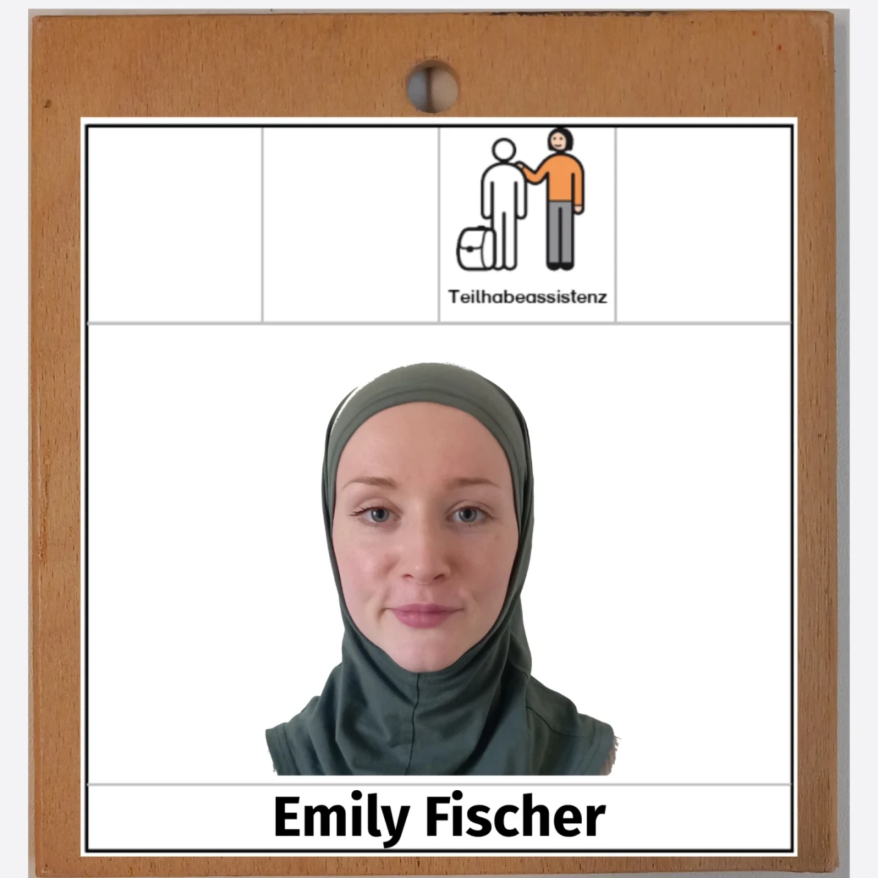 Portr&auml;tfoto einer Frau mit hellgr&uuml;nem Kopftuch vor wei&szlig;em Hintergrund. &Uuml;ber ihrem Bild ein Logo mit zwei gezeichneten Figuren, davon eine Person im Rollstuhl; rechts daneben der Text &bdquo;Teilhabeassistenz&ldquo;. Hintergrund ein brauner Holzrahmen. Darunter steht der Name &bdquo;Emily Fischer&ldquo;.