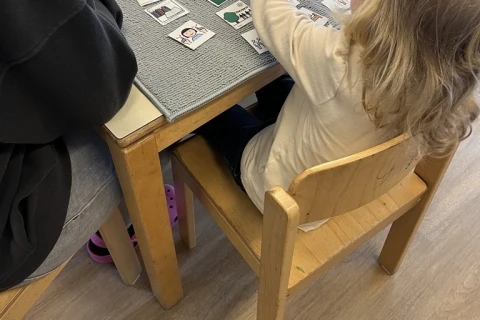 Kind mit blondem Haar in wei&szlig;em Langarmshirt sitzt auf einem Holzstuhl an einem Tisch und ordnet K&auml;rtchen auf grauem Filz. Links sitzt eine Person in dunkler Kleidung. Auf dem Tisch liegen Papierunterlagen und eine weitere Tafel mit Bildsymbolen. Laminierter Holzfu&szlig;boden im Hintergrund.