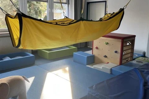 Gelber H&auml;ngetuch-Sitz, aufgeh&auml;ngt an vier Seilen an der Decke in einem Raum mit Fenstern und Tageslicht. Darunter blaue und gr&uuml;ne Polstermatten, links ein Polstertier in Elefantenform, rechts ein Kletterw&uuml;rfel mit bunten Griffen und weitere Matten.