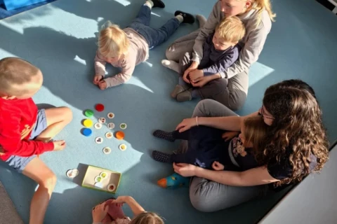 F&uuml;nf Kinder und ein Erwachsener auf einer hellblauen Spielmatte in einem Klassenraum. Auf der Matte liegen bunte Spielchips, sortiert in Farben, und eine offene Box mit weiteren Spielmaterialien. Im Hintergrund stehen eine blaue Plastikbox und eine Holzbox mit Bausteinen. Licht- und Schatteneffekte durch Sonneneinfall.
