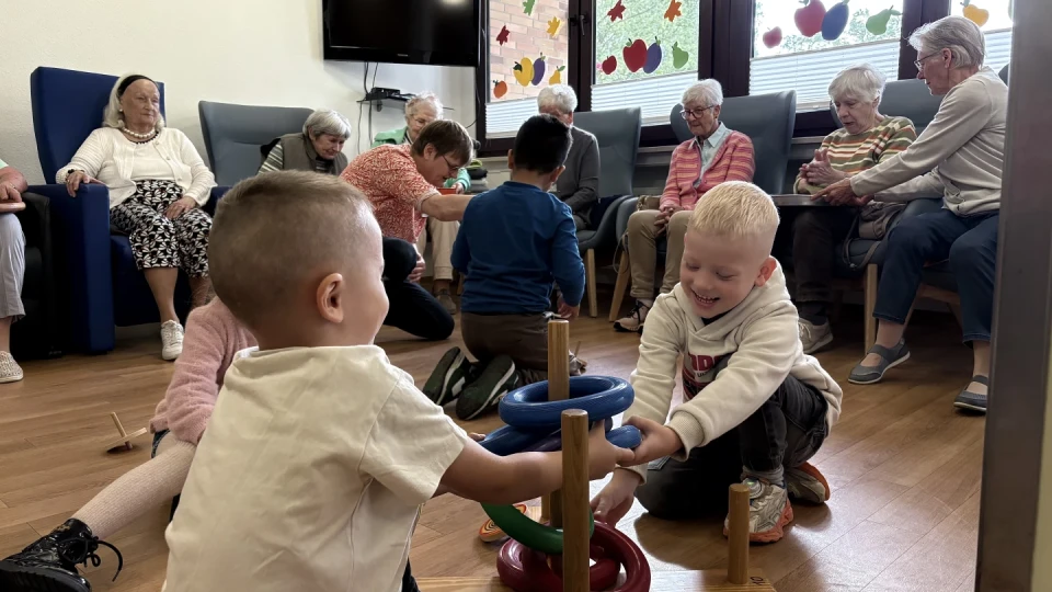 Kinder spielen mit bunten Ringen, die sie auf Holzstangen stecken, auf einem Holzboden. Dahinter sitzen &auml;ltere Menschen auf St&uuml;hlen, in Gespr&auml;ch vertieft. Fenster mit herbstlichen Dekorationen wie Bl&auml;ttern und &Auml;pfeln im Hintergrund, daneben ein Fernseher an der Wand.
