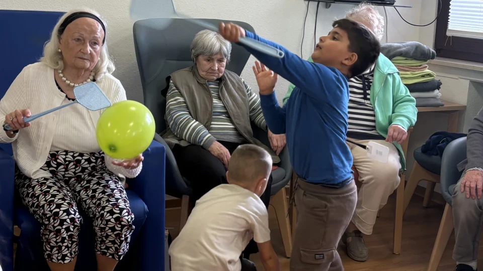 &Auml;ltere Menschen und Kinder spielen mit bunten Luftballons in einem Raum mit Holzboden. Eine &auml;ltere Frau h&auml;lt einen blauen Schl&auml;ger, ein Junge im blauen Shirt schl&auml;gt einen Ballon, w&auml;hrend ein kleinerer Junge sich b&uuml;ckt. Im Hintergrund sitzen weitere Personen auf St&uuml;hlen, an der Wand ein Fernseher.