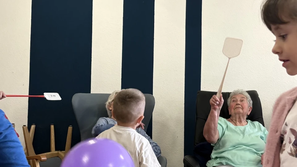 &Auml;ltere Frau in einem hellgr&uuml;nen Oberteil und beiger Hose sitzt auf einem dunklen Stuhl und h&auml;lt eine Fliegenklatsche in der Hand. Kinder, darunter ein Junge mit wei&szlig;em T-Shirt und eine Ballon spielende Gruppe. Hintergrund mit cremefarbener Wand und blauen, vertikalen Streifen.