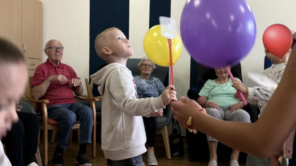 Kinder spielen mit bunten Luftballons und gelben Plastik-Schl&auml;gern in einem Raum mit Holzboden. Im Hintergrund sitzen &auml;ltere Erwachsene auf Holzst&uuml;hlen entlang der W&auml;nde vor dunkelblauen Streifen an einer wei&szlig;en Wand und beobachten das Spiel.