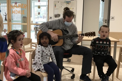 Mehrere Kinder sitzen in einem Stuhlkreis und singen. Ein Mann psielt dazu Gitarre