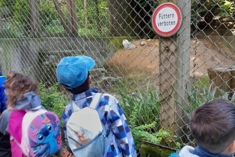 Kinder mit bunten Rucks&auml;cken und Jacken stehen vor einem Metallzaun mit einem Schild, das "F&uuml;ttern verboten" zeigt. Hinter dem Zaun ist eine sandige Fl&auml;che, Vegetation und ein Tiergehege mit Holzkonstruktionen sichtbar.