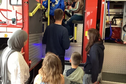 Mehrere Menschen, darunter Erwachsene und Kinder, stehen neben einem ge&ouml;ffneten Feuerwehrauto in einer Halle. Einige Kinder klettern in das rote Feuerwehrauto mit gelben Haltestangen und hell erleuchtetem Innenraum. Rechts sind Ger&auml;tef&auml;cher des Fahrzeugs sichtbar, darin zusammengerollte Schl&auml;uche. Der Boden ist aus rotem Fliesenbelag mit gelben Markierungen.