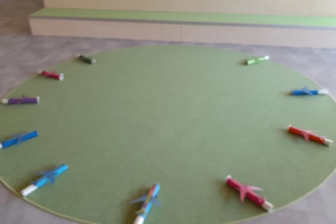 Gr&uuml;ner, kreisf&ouml;rmiger Teppich auf einem grau gefliesten Boden, umgeben von kleinen Spielzeugfliegern in verschiedenen Farben wie Blau, Rot, Gr&uuml;n, Lila und Pink. Im Hintergrund eine flache B&uuml;hne mit gr&uuml;nem Teppichbelag, darauf mehrere gestapelte Kisten, eine gepolsterte Bank und Schultaschen. W&auml;nde in hellem Beige-Ton, Fenster auf der rechten Seite.