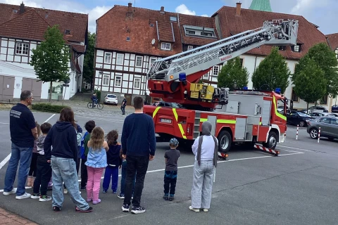 Gruppe von Erwachsenen und Kindern auf einem Platz, die ein rotes Feuerwehrfahrzeug mit ausgefahrener Drehleiter betrachten. Im Hintergrund Fachwerkh&auml;user, B&auml;ume und ein Kirchturm mit gr&uuml;ner Spitze. Der Himmel ist bew&ouml;lkt mit blauen und wei&szlig;en Abschnitten.