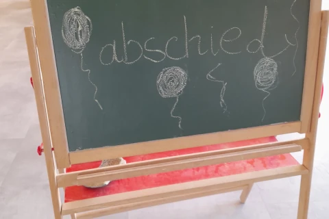 Gr&uuml;ne Tafel auf einem Holzgestell mit der Kreideaufschrift &bdquo;Schulkinderabschied&ldquo;. Umgeben von f&uuml;nf gezeichneten Ballons und geschwungenen Linien. Darunter liegt ein roter Tafelschwamm auf einem roten Filzunterlageteil. Im Hintergrund unscharfer grauer Boden.