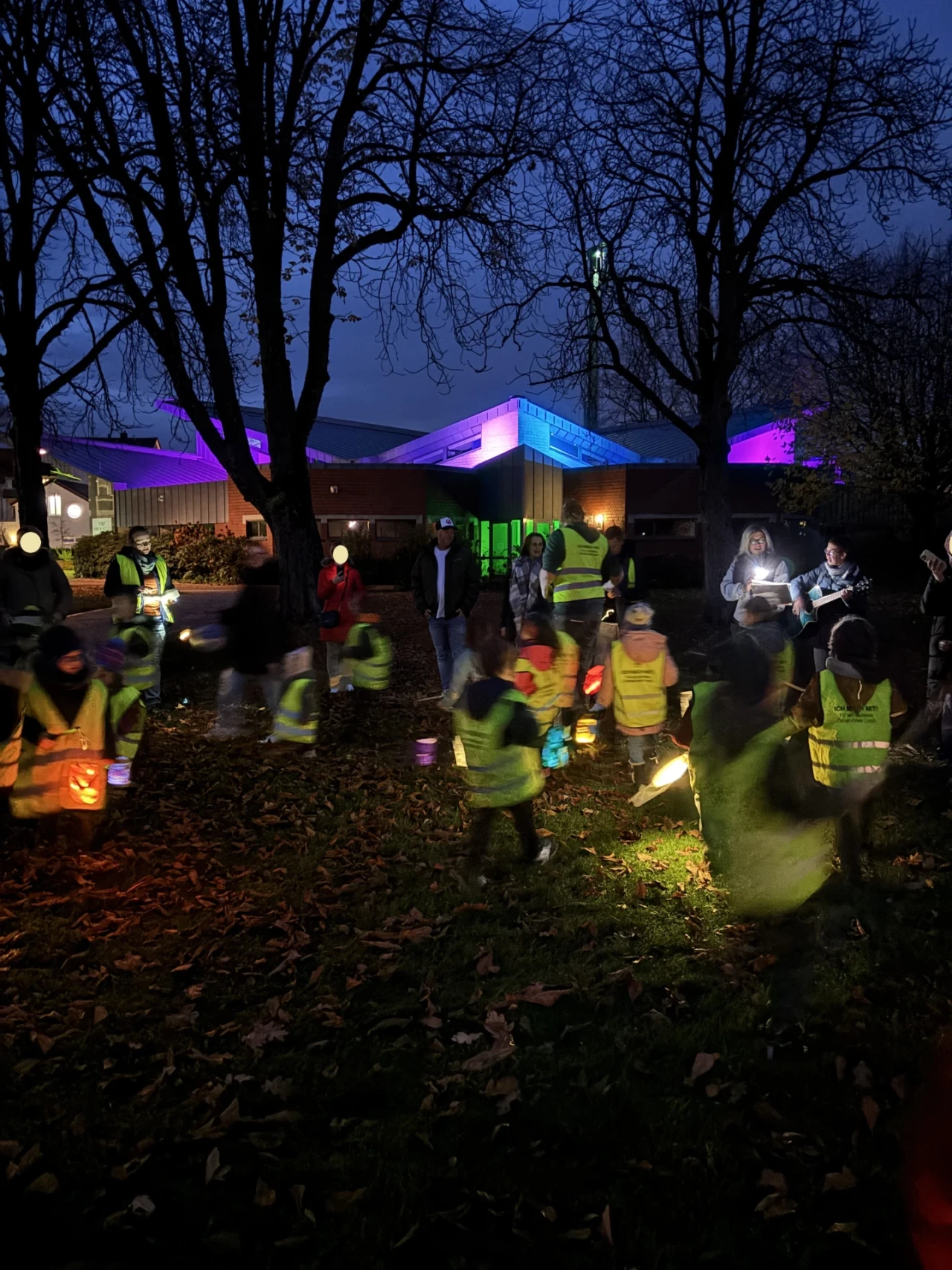 Kinder in reflektierenden gelben Westen mit bunten Laternen versammelt auf einer Wiese voller Herbstlaub; dahinter stehen Erwachsene, teils musizierend. Im Hintergrund ein Geb&auml;ude mit beleuchteten W&auml;nden in Blau, Lila und Gr&uuml;n, umgeben von kahlen B&auml;umen im Dunkeln.