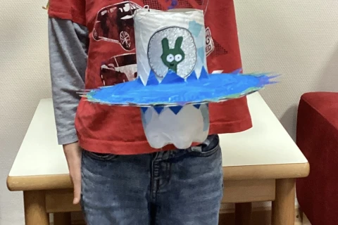 Ein Kind in einem roten Langarmshirt mit Autoaufdrucken und blauer Jeans h&auml;lt eine selbstgebastelte Laterne. Die Laterne besteht aus einer wei&szlig;en, zylinderf&ouml;rmigen Basis mit einem gr&uuml;nen Hasenmotiv und einer breiten blauen Scheibe mit gezackten R&auml;ndern. Die Laterne ist an einem Drahtb&uuml;gel befestigt. Im Hintergrund sind ein Holztisch, wei&szlig;e W&auml;nde und ein rotes Sofa sichtbar.
