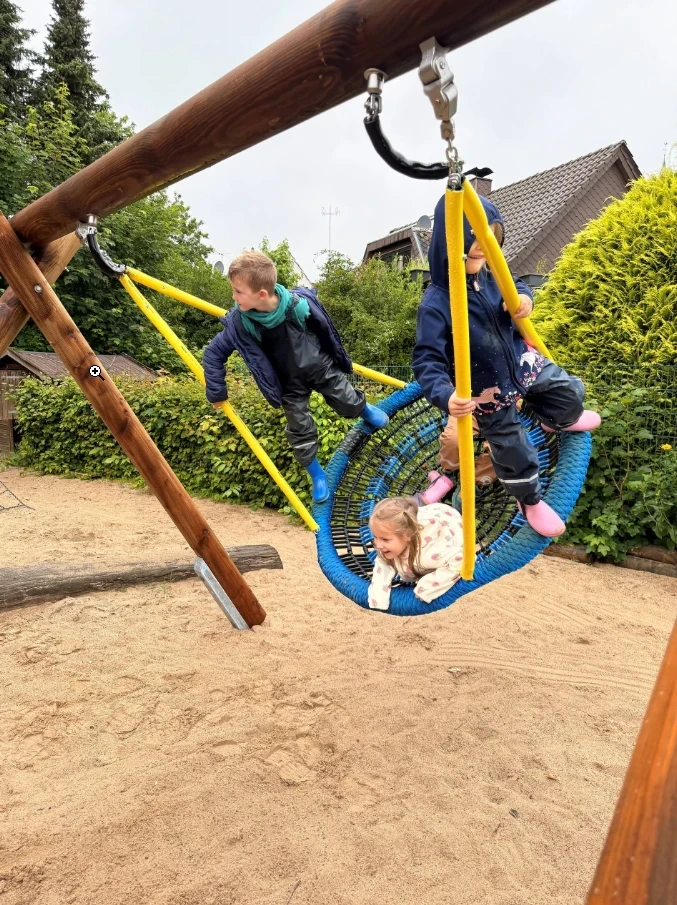 Drei Kinder spielen auf einer blauen Netzschaukel, die an einem Holzgestell mit gelben Haltestangen h&auml;ngt. Zwei Kinder sitzen, ein weiteres Kind liegt auf der Schaukel. Der Sandboden und B&uuml;sche sowie ein Haus mit Schr&auml;gdach sind im Hintergrund sichtbar.