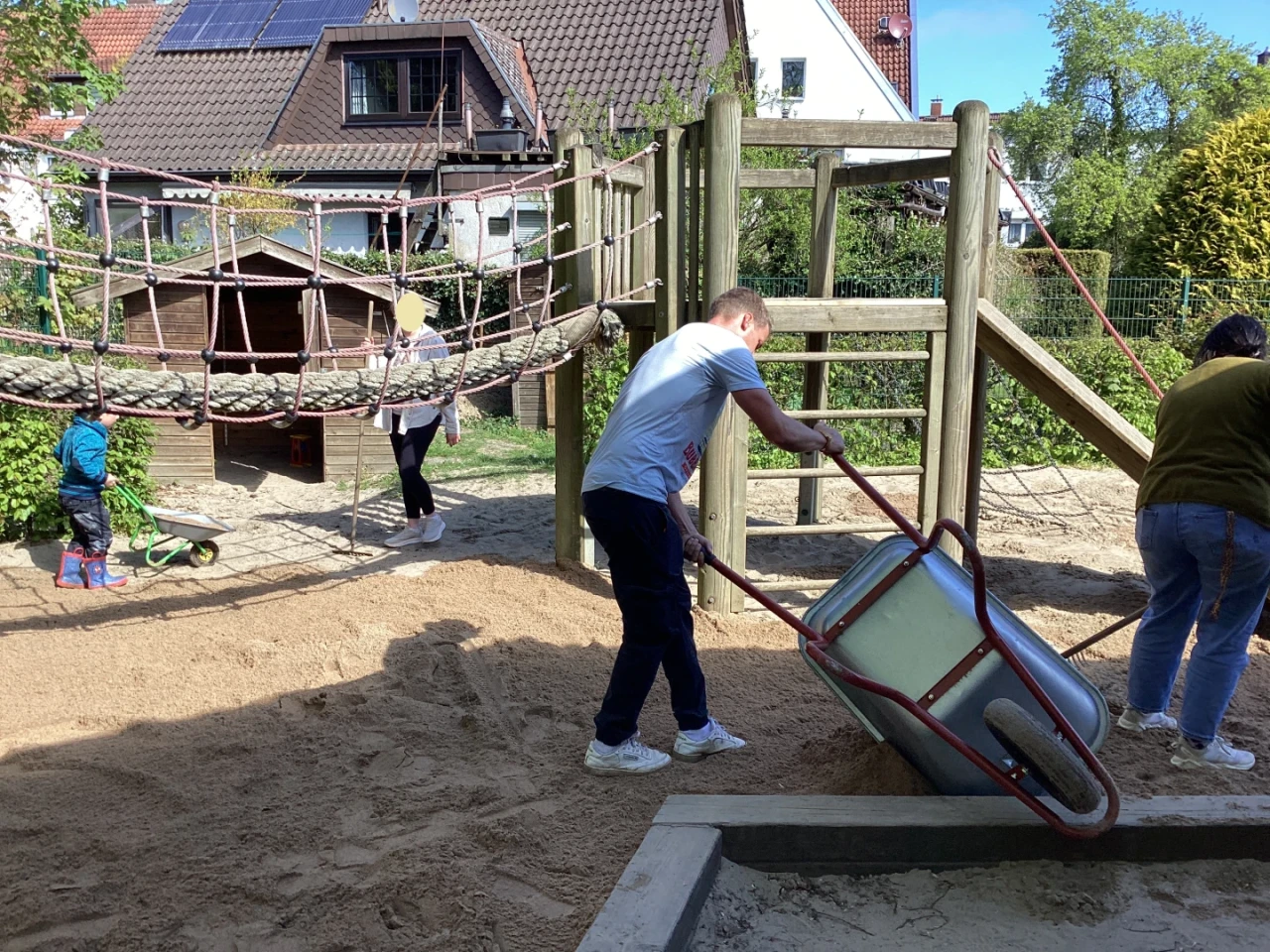Mann mit wei&szlig;em T-Shirt, der Sand mit einer Schubkarre auf einen Sandspielplatz transportiert. Rechts und links zwei weitere Erwachsene, im Hintergrund Holzspielger&auml;te mit Kletternetz und eine Gartenh&uuml;tte, umgeben von Wohnh&auml;usern und B&auml;umen. Zwei Kinder spielen links in der N&auml;he eines Sandkastens.