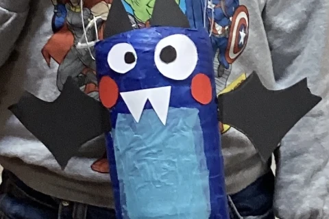 Selbstgebastelte Laterne in Form einer blauen Fledermaus aus einer Plastikflasche, mit gro&szlig;en wei&szlig;en Augen, roten Wangen, spitzen wei&szlig;en Z&auml;hnen und schwarzen Kartonfl&uuml;geln. Gehalten von einer Hand, Person tr&auml;gt ein graues Sweatshirt mit Comicfiguren und eine blaue Jeans.