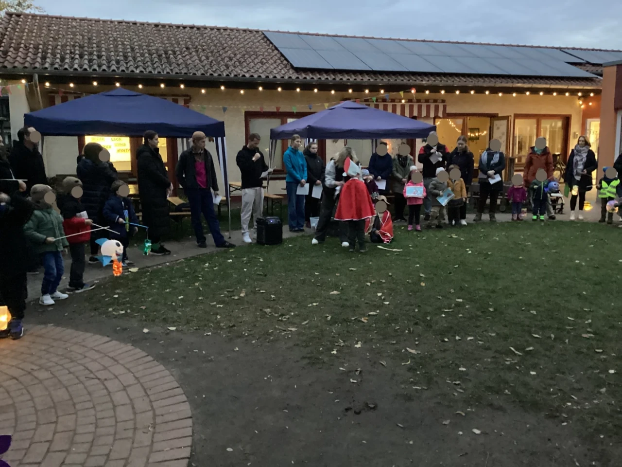Menschenansammlung im Freien vor einem Geb&auml;ude mit Ziegeldach und Solarpanels am fr&uuml;hen Abend. Erwachsene und Kinder, einige mit bunten Laternen, bilden einen Kreis auf einem Grasplatz. Zwei Pavillons und kleine Lichterketten sind sichtbar. Ein Mann mit rotem Mantel und Stab steht in der Mitte.
