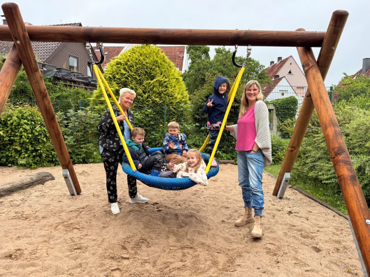 Holz-Schaukelsitz mit Netzgeflecht auf einem Sandspielplatz, mehrere Kinder sitzen darin, zwei Erwachsene stehen daneben. Hintergrund mit gr&uuml;nen B&uuml;schen, B&auml;umen und mehreren H&auml;usern mit roten und wei&szlig;en Fassaden.