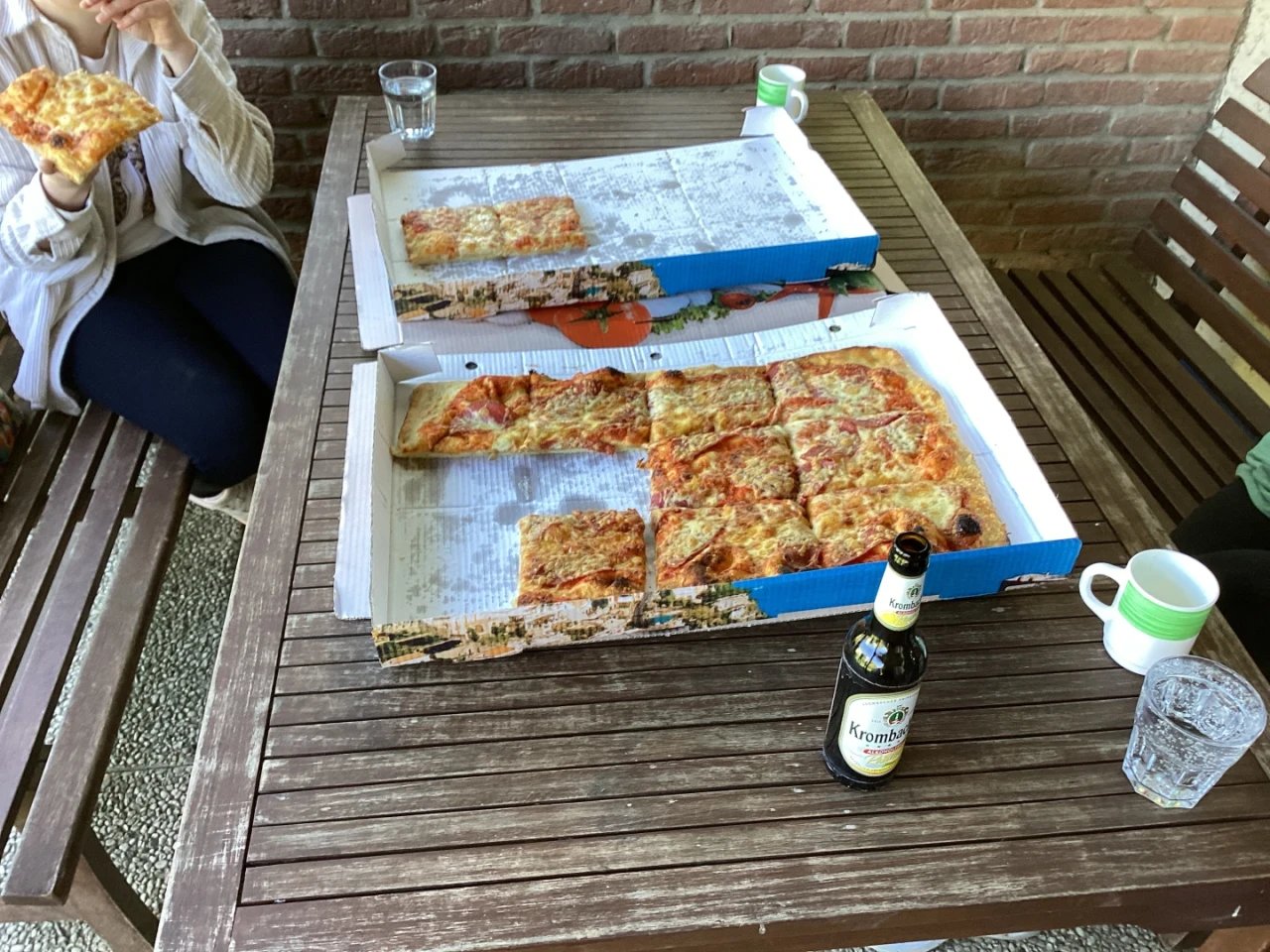 Zwei ge&ouml;ffnete Pizzakartons auf einem braunen Holztisch, eine Pizza fast vollst&auml;ndig, die andere teils gegessen. Eine Person h&auml;lt ein Pizzast&uuml;ck, daneben gr&uuml;ne Tassen und Gl&auml;ser mit Getr&auml;nken. Eine Bierflasche "Krombacher" vorne rechts. Backsteinwand im Hintergrund.