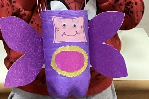 Person h&auml;lt eine selbstgebastelte Laterne in Form eines lila Schmetterlings aus Papier. Die Laterne hat zwei gro&szlig;e seitliche Fl&uuml;gel, ein Gesicht mit Wackelaugen und einem aufgemalten Mund auf rosa Hintergrund, sowie ein pink-gelbes, rundes Dekorelement unten. Hintergrund: Tisch und helle Wand.