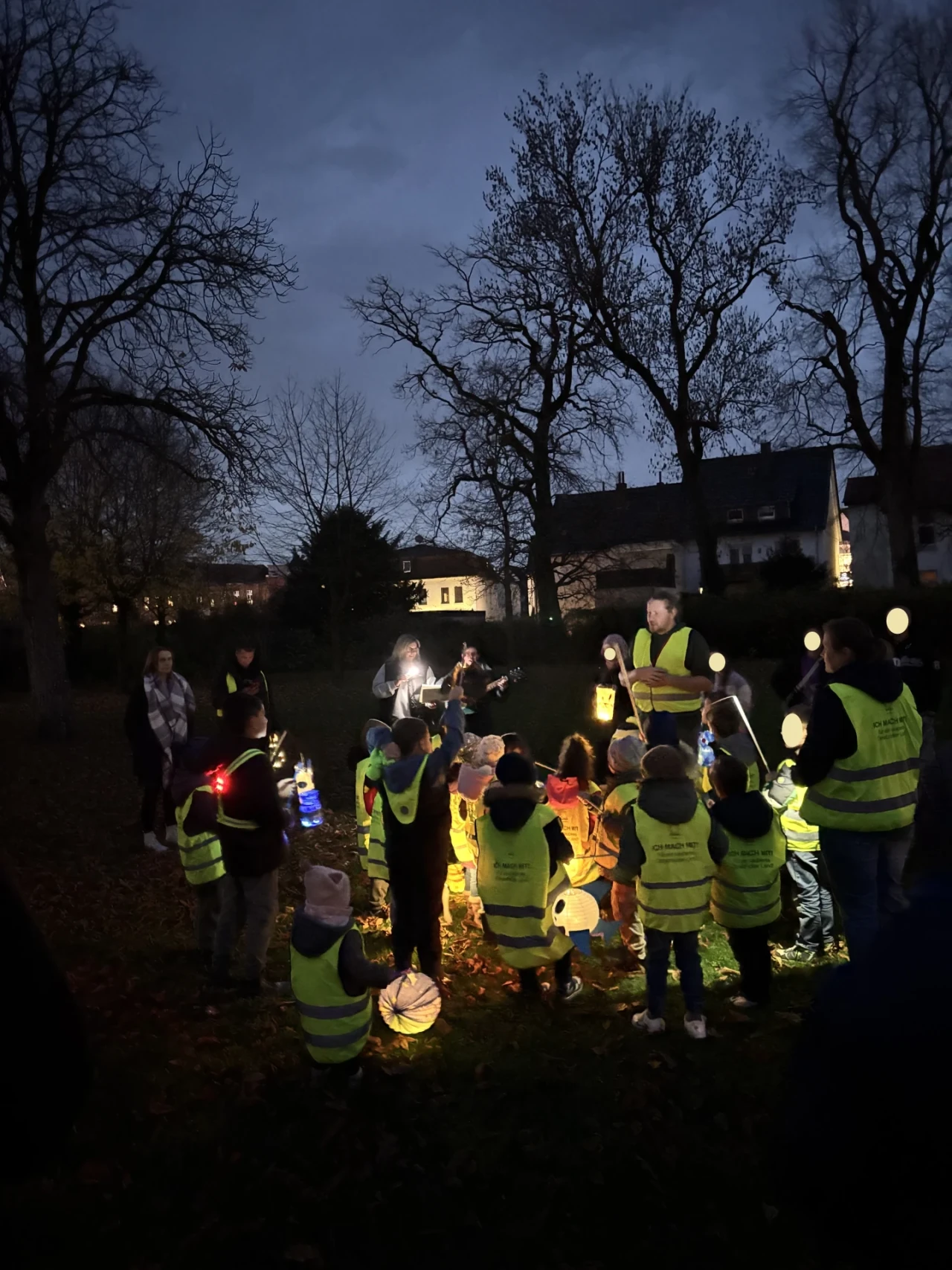 Gruppe von Kindern in gelben Sicherheitswesten, die beleuchtete Laternen tragen, begleitet von Erwachsenen, ebenfalls in Sicherheitswesten. Sie stehen auf einer Wiese mit kahlen B&auml;umen im Hintergrund, der Himmel ist dunkelblau. Wohnh&auml;user mit teils beleuchteten Fenstern im Hintergrund. Ein Erwachsener h&auml;lt eine leuchtende Laterne hoch.