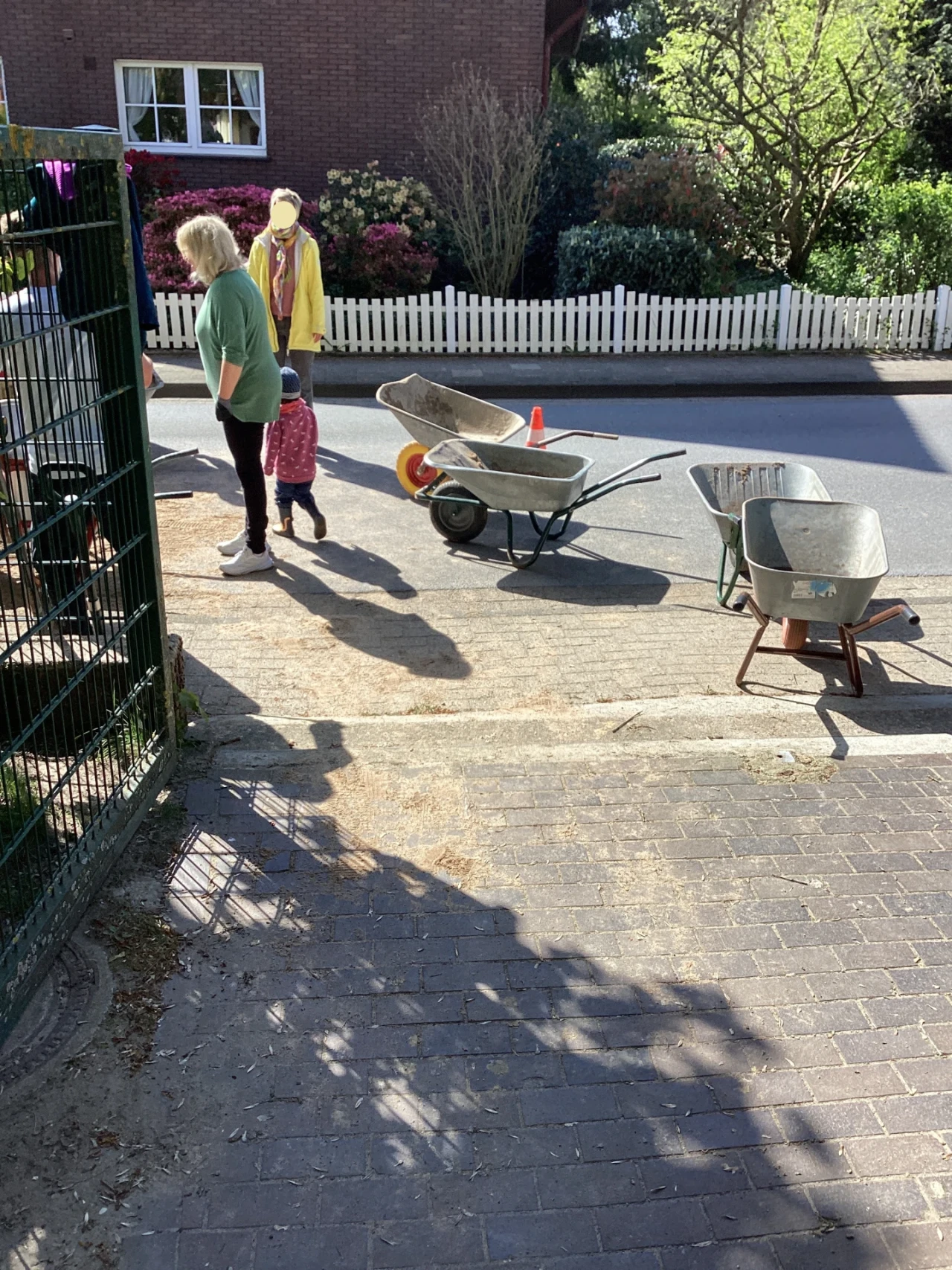 Zwei Schubkarren und eine Kinderkarre stehen auf einem gepflasterten Weg. Zwei Erwachsene und ein Kind in pinker Kleidung sind sichtbar. Hinter ihnen eine Stra&szlig;e, ein wei&szlig;er Gartenzaun, Str&auml;ucher und ein Backsteinhaus mit Fenstern.