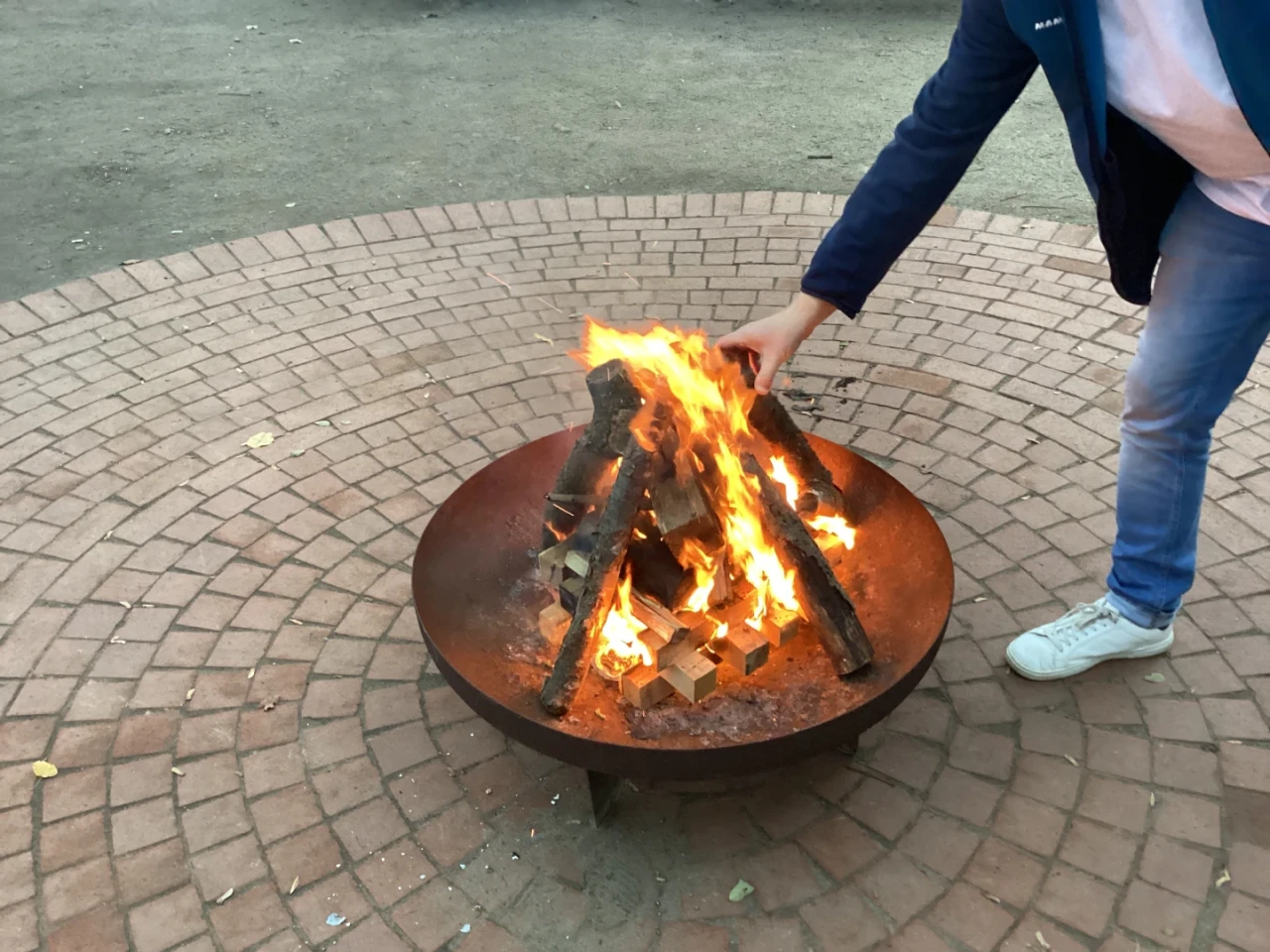 Brennendes Lagerfeuer in einer runden, flachen Metallschale, umgeben von einem gepflasterten Boden aus r&ouml;tlich-beigen Steinen. Eine Person mit wei&szlig;em Schuh, blauer Jeans und dunkelblauer Jacke positioniert ein Holzst&uuml;ck mit der rechten Hand ins Feuer.