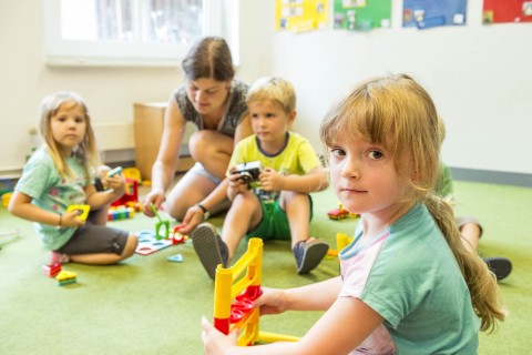 Mehrere Kinder und eine Betreuerin spielen zusammen im Kindergartenraum