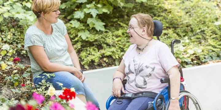 Zwei Frauen in einem Gartenbereich mit Blumen und gr&uuml;nen Str&auml;uchern. Links sitzt eine Frau mit kurz geschnittenem, blondem Haar aufrecht und tr&auml;gt eine hellgr&uuml;ne Bluse sowie blaue Jeans. Rechts sitzt eine Frau mit kurzen, r&ouml;tlichen Haaren in einem blauen Rollstuhl. Sie tr&auml;gt ein rosafarbenes T-Shirt mit Schmetterlingsmotiv und spricht l&auml;chelnd mit der Frau. Im Vordergrund bunte Blumen, im Hintergrund dichte, gr&uuml;ne Pflanzen.