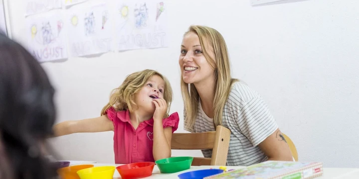 Eine Frau mit hellblondem Haar im gestreiften T-Shirt und ein kleines M&auml;dchen mit blonden Haaren im pinken &auml;rmellosen Oberteil sitzen an einem Tisch. Auf dem Tisch stehen bunte Schalen, Stifte und Bastelmaterial. Im Hintergrund wei&szlig;e Wand mit Kinderzeichnungen und Wort "August".