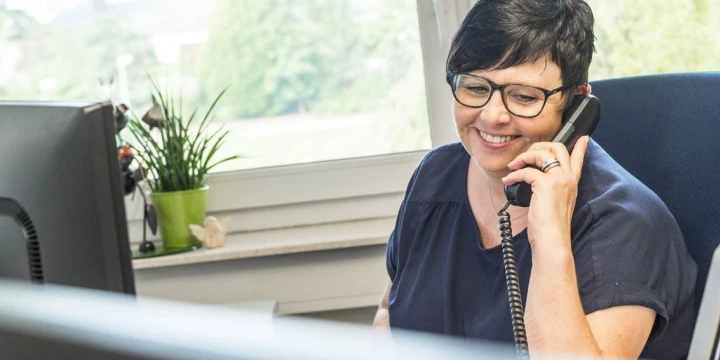 Person in dunkelblauer Kleidung sitzt an einem Schreibtisch und h&auml;lt ein schwarzes Telefon am Ohr. Im Hintergrund ein Fenster mit Blick auf gr&uuml;ne Vegetation, daneben ein gr&uuml;ner Blumentopf mit Pflanze und ein Monitor am linken Bildrand.