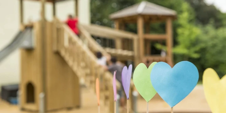 Bunte Papierherzen auf Holzst&auml;ben im Vordergrund, in Blau, Gr&uuml;n, Gelb und Lila. Im Hintergrund ein h&ouml;lzerner Spielplatz mit Kletterger&uuml;st, Rutsche und zwei kleinen Pavillons, umgeben von gr&uuml;nem Laub. Einige Kinder sind am Spielger&auml;t.