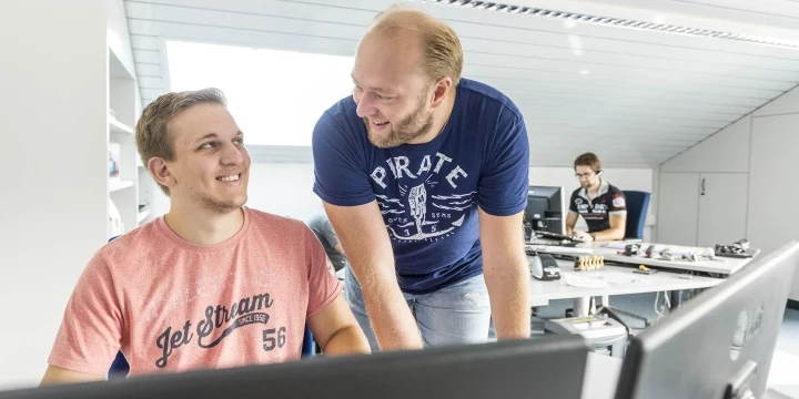 Zwei M&auml;nner in einem modernen B&uuml;ro, der linke tr&auml;gt ein rotes T-Shirt mit der Aufschrift "Jet Stream 56" und sitzt vor einem Monitor l&auml;chelnd. Der rechte, in einem blauen T-Shirt mit "Pirate"-Schriftzug, beugt sich zu ihm hin&uuml;ber und l&auml;chelt. Im Hintergrund sitzt ein weiterer Mann an einem Schreibtisch mit Computern und Werkzeug. Licht durch gro&szlig;e Fenster.