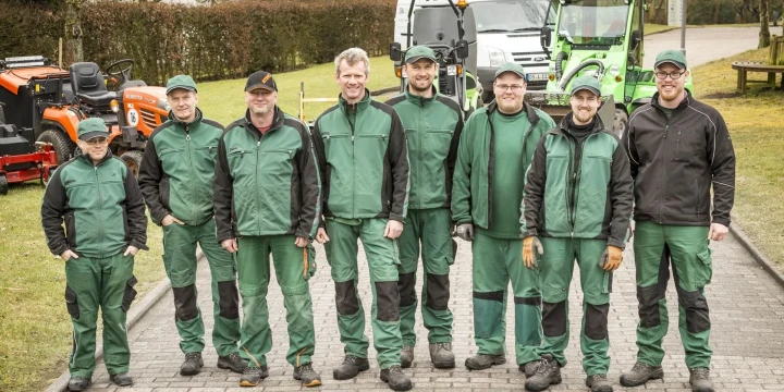 Acht M&auml;nner in gr&uuml;ner Arbeitskleidung stehen auf einer gepflasterten Stra&szlig;e nebeneinander. Im Hintergrund sind ein wei&szlig;er Transporter, ein gr&uuml;nes und ein orangefarbenes Arbeitsfahrzeug sowie ein Zaun, Rasen und H&auml;user mit roten D&auml;chern sichtbar.