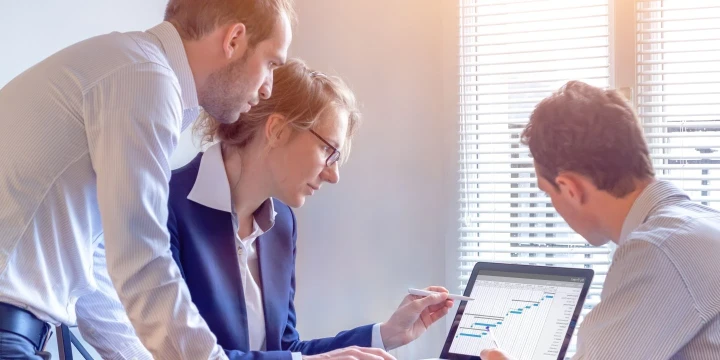 Drei Personen in Business-Kleidung arbeiten an einem Tisch. Eine Frau in einem dunkelblauen Blazer mit Brille zeigt mit einem Stift auf ein Laptop-Bildschirm mit einem Gantt-Diagramm. Zwei M&auml;nner, einer links stehend, der andere rechts sitzend, beobachten den Bildschirm konzentriert. Licht scheint durch Jalousien rechts im Bild.