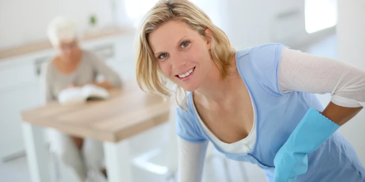 Blonde Frau in hellem blauem Shirt und wei&szlig;en &Auml;rmeln l&auml;chelt, w&auml;hrend sie mit blauen Gummihandschuhen arbeitet. Im Hintergrund sitzt eine &auml;ltere Frau mit kurzen wei&szlig;en Haaren an einem Holztisch und liest ein Buch. Der Raum ist hell und minimalistisch eingerichtet.