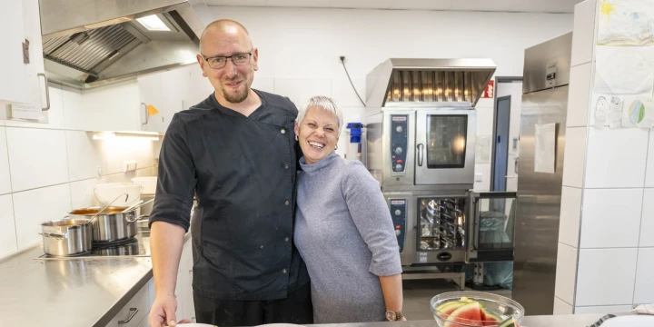 Mann in schwarzer Kochkleidung und Frau in grauem Pullover stehen l&auml;chelnd in einer Gro&szlig;k&uuml;che vor einer Edelstahl-Arbeitsfl&auml;che. Auf der Arbeitsfl&auml;che liegen vier Teller mit aufgeschnittenem Gem&uuml;se wie Gurken, Paprika und Karotten sowie eine Sch&uuml;ssel mit Melonenst&uuml;cken. Im Hintergrund K&uuml;chenger&auml;te wie ein Dampfgarer und T&ouml;pfe.