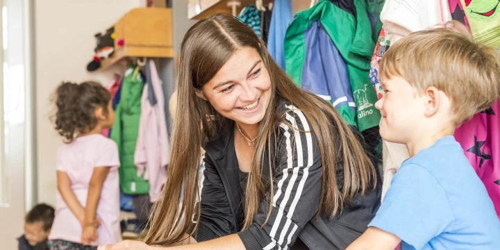 Frau mit braunen, langen Haaren, schwarzer Jacke mit wei&szlig;en Streifen an den &Auml;rmeln, sitzt l&auml;chelnd auf einer Bank und unterh&auml;lt sich mit einem Jungen in blauem T-Shirt. Im Hintergrund Garderobe mit bunt sortierten Kinderjacken und zwei weiteren Kindern bei der Garderobe.
