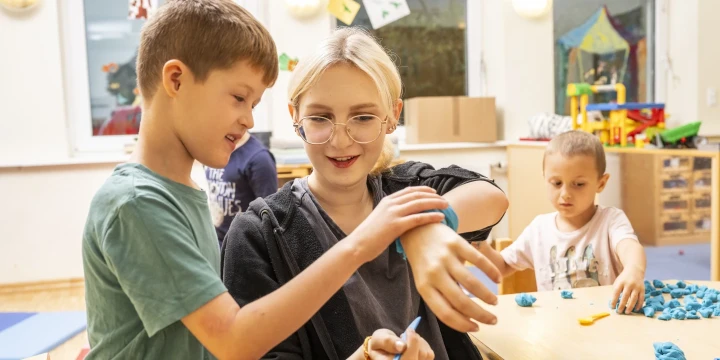 Junge mit gr&uuml;nem T-Shirt und Brille bindet einer blonden jungen Frau eine blaue Tonfigur um das Handgelenk. Rechts spielt ein Kind mit heller Kleidung mit blauem Knetmaterial an einem Holztisch. Im Hintergrund Fenster, eine gelbe Wanddekoration und Spielzeug.