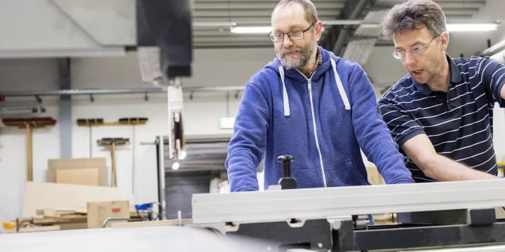 Zwei M&auml;nner, einer mit blauer Kapuzenjacke und einer mit dunklem gestreiften Polohemd, stehen an einer gro&szlig;en Maschine in einer Werkstatt. Im Hintergrund sind Regale, Kartons und Holzbretter sichtbar, die von einer Betonwand und einer Decke mit Metalltr&auml;gern umgeben sind.